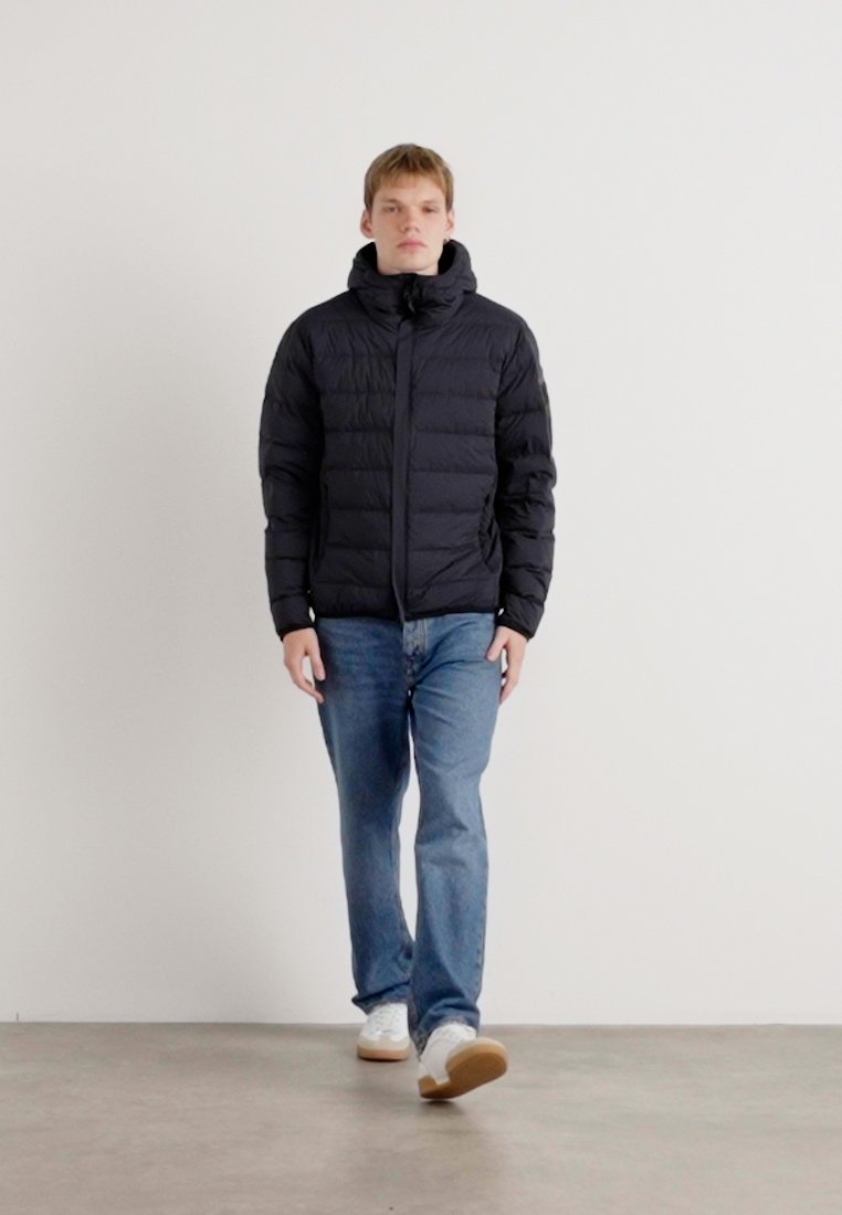 Doudoune noire avec capuche, design matelassé, fermeture éclair à l'avant, portée sur un jean bleu et des baskets blanches, se tenant sur un fond neutre.