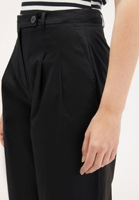 Pantalons plissés noirs avec une texture lisse, dotés d'une fermeture à bouton et de poches latérales. Détail d'une main reposant sur le tissu.