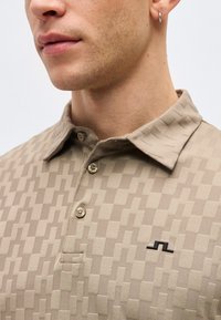 Κοντινό πλάνο ενός άνδρα που φοράει μπεζ πουκάμισο πόλο με σχέδια, κουμπιά και ένα μικρό μαύρο λογότυπο στο στήθος, καθώς και ένα μικρό σκουλαρίκι κρίκο.