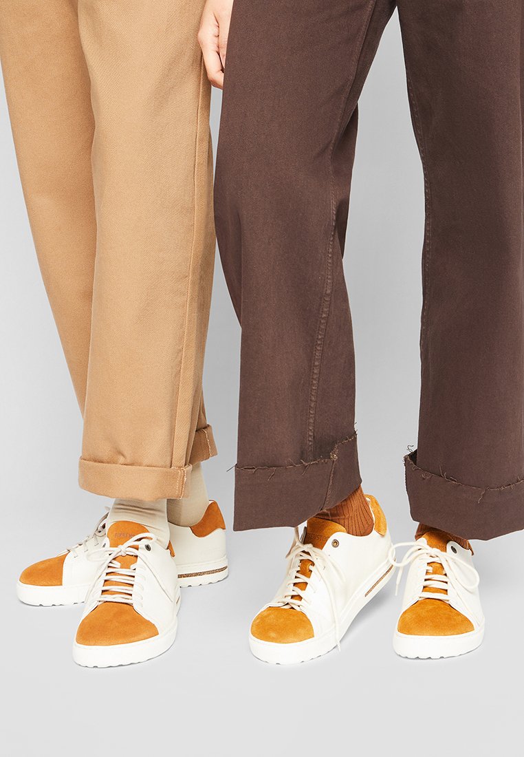 Weiße und orange Sneaker mit einer Veloursleder-Zehenschutzkappe und Zunge, kombiniert mit beigen und braunen, umgeschlagenen Hosen mit fransigen Säumen.