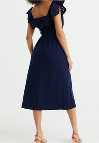 Navyblauwe jurk met een smocked lijfje en off-the-shoulder mouwen met kantafwerking. Vloeiende midi-rok, zachte textuur en relaxte pasvorm.