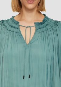 Türkise, gefaltete Bluse mit einem hohen, gerafften Kragen und einem V-Ausschnitt. Verfügt über eine Kordelzugverschluss mit schwarzen Aglets. Glatte Stoffstruktur.