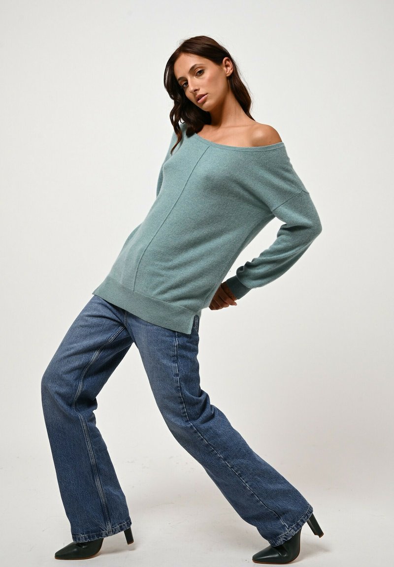 Helles blauer, schulterfreier Pullover mit strukturiertem Strick, kombiniert mit hoch taillierten, weit geschnittenen dunklen Jeans und schwarzen Absatzschuhen.