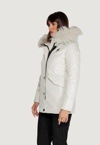 Parka bianca isolata con cappuccio in pelliccia sintetica, caratterizzata da chiusura con zip, tasche laterali e dettagli neri, texture liscia e vestibilità sartoriale.