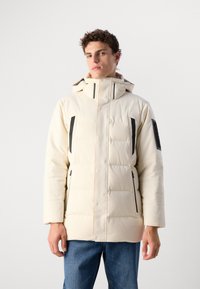 Helle beigefarbene, gepolsterte Jacke mit schwarzen Akzenten, hohem Kragen, Reißverschlusstaschen und Kapuze. Texturierte Oberfläche mit matter Optik.