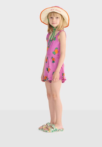 Jeune fille portant une combinaison-short rose avec des imprimés de carottes, un chapeau à large bord avec des liens verts, et des sandales à motif floral, debout de profil.