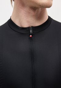 Uomo che indossa una camicia nera con cerniera e collo rotondo, cerniera completamente chiusa e una piccola linguetta rossa della cerniera visibile al collo.