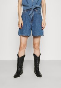 Helle blaue Jeansshorts mit lockerer Passform und genähten Säumen, ergänzt durch schwarze Cowboystiefel mit geprägten Details und spitz zulaufenden Zehen.