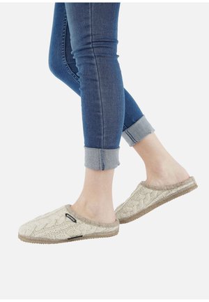 Knitted beige slippers with a textured pattern and light tan sole. Worn with rolled-up blue denim jeans, showcasing the slippers' side view.