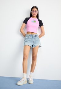 Ung kvinna iförd en rosa och svart kortärmad t-shirt med grafik, slitna jeansshorts, vita strumpor och pastellfärgade sneakers, stående mot en vit vägg.