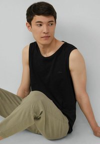 Schwarzes ärmelloses Tanktop aus Baumwolle, kombiniert mit hellolivgrünen Hosen. Einfaches Design mit kleinem Logo auf der Brust.