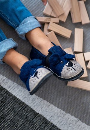 Kinderfüße in blauen Hausschuhen mit Hundemotiv, umgeben von zerstreuten Holzbauklötzen auf einem Teppichboden.