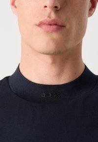 Navy crewneck t-skjorte laget av glatt stoff med en teksturert krage, med en hevet svart logo ved kragen.