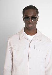 Lichtbeige jas met een knoopsluiting aan de voorkant en twee borstzakken, gedragen over een wit T-shirt. Zwarte rechthoekige zonnebril maakt de look compleet.
