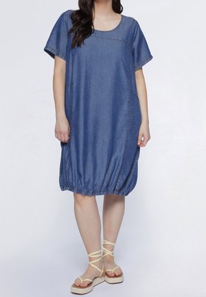 Kvinde iført løs blå denim kjole med korte ærmer og samlet kant, kombineret med beige kilehælssandaler med anklerem.