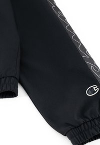 Pantalons de sport noirs avec des poignets élastiques, featuring un panneau latéral texturé avec un motif de vagues blanches et un petit logo brodé.