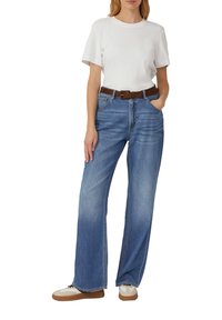 T-shirt blanc à manches courtes, jean bleu à jambes larges avec un délavage léger, ceinture marron et baskets beiges avec des lacets noirs.