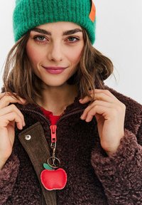 Mladá žena v zelené pletené čepici a hnědé chlupaté bundě s klíčenkou v podobě červené jablka na zipu, jemně se usmívající do kamery.