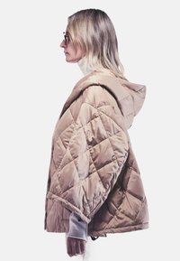 Gequilte beige pufferjack met een capuchon, met een losse pasvorm, ruitpatroon en een zachte glansafwerking. Draagt over een lichte turtleneck.