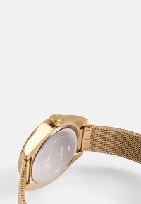 Gold-tone watch with a mesh band, round case, and prominent crown. Back features engraved text and a smooth metallic surface.