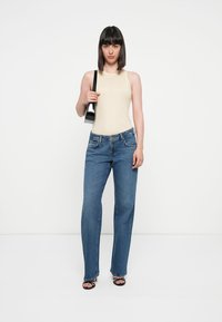 Beige ermeløs topp, høyt liv blå jeans, svart håndveske og svarte sandaler med stropper. Modellen står rett opp mot en hvit bakgrunn.