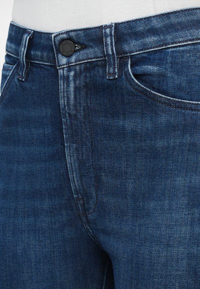 Primo piano della vita di jeans in denim blu scuro con bottone e tasca frontale, indossati con una maglietta bianca infilata dentro.
