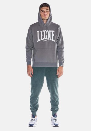 Jeune homme portant un sweat à capuche gris avec "LEONE 1947," jogging vert foncé et baskets blanches, debout face à l'avant avec les mains détendues.