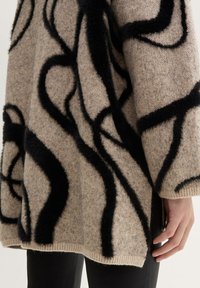 Beige neulepaita, jossa on mustia, teksturoituja abstrakteja kuvioita. Löysä malli, leveä pääntie ja ribbireunus.