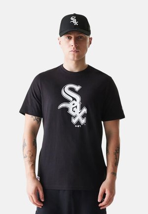 Mies, joka pitää mustaa Chicago White Sox -lippistä ja vastaavaa mustaa t-paitaa, jossa on suuri valkoinen Sox-logo, seisoo rentoutunein käsin sivuillaan.