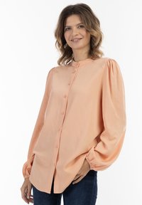 Persikofärgad skjorta med knappar och volangärmar, slät textur och rundad krage. Bärs med mörka jeans.