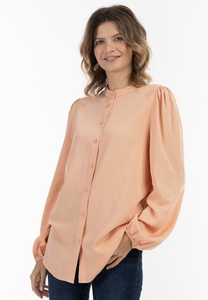 Ferskenfarvet skjorte med knapper, fyldige og puffede ærmer, glat tekstur og rund krave. Båret med mørke jeans.