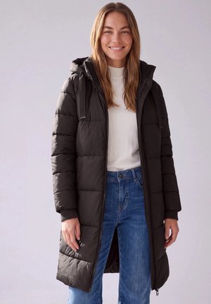 Femme souriante portant une longue doudoune noire à capuche, un pull blanc et un jean bleu sur un fond clair uni.