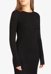 Robe pull noire en maille côtelée avec des manches longues, silhouette ajustée et trous pour les pouces. Motif texturé sur l'ensemble. Encolure ras du cou.