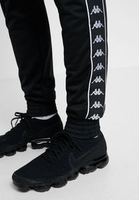 Svarta Kappa-träningsoveraller har en vit sidostrand med upprepade logotypmönster. I kombination med svarta Nike-skor och ribbade anklar.