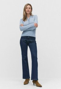 Kvinna som står med armarna i kors iklädd en ljusblå långärmad topp, mörkblå jeans och bruna klackankelboots mot en enfärgad bakgrund.