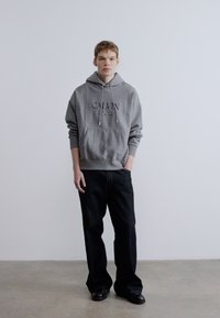 Szürke kapucnis pulóver "CALVIN" felirattal, elülső zsebbel és húzózsinóros kapucnival; sötét farmernadrággal és fekete cipővel párosítva, semleges háttéren állva.