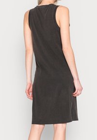 Robe noire sans manches, longueur genoux, avec des bords bruts, portée par une personne vue de dos sur un fond uni.