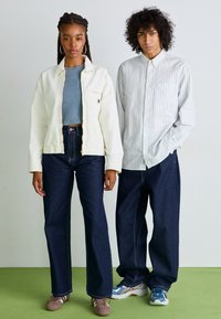To modeller står side om side. Kvinnen har på seg en kremfarget zip-jakke, en blå kort topp og mørke vide jeans. Mannen har på seg en stripet skjorte med knapper og baggy mørke jeans, sammen med sporty sko.