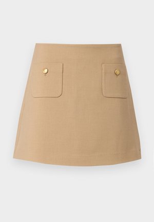 Mini-jupe en lin beige avec deux poches avant, chacune dotée d'un bouton doré. Design simple et coupe droite avec une fente sur le côté.