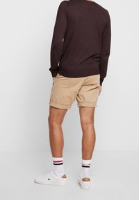 Pantaloni beige con risvolti, abbinati a una camicia a maniche lunghe marrone scuro. Sneakers bianche con dettagli color marrone chiaro e calzini a righe.