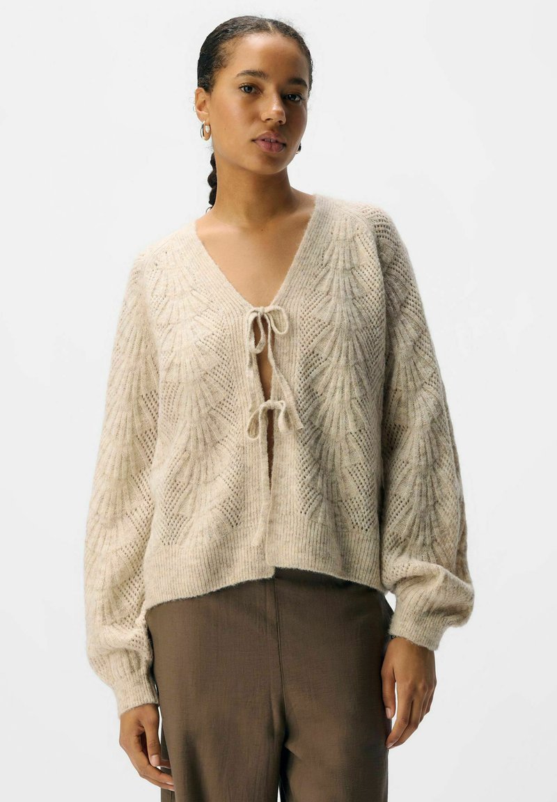 Ljusbeige stickad cardigan med V-ringning, knytframslutning och texturerat lövmönster. Långa, voluminösa ärmar och croppad nederkant.