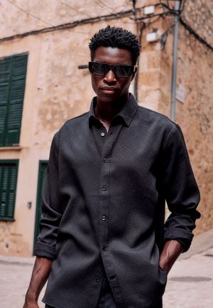 Man with short curly hair wearing black sunglasses and a black long-sleeve shirt standing on a stone-paved street with shuttered windows.