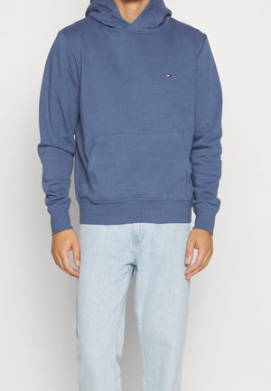 Felpa blu in morbido tessuto, con tasca frontale a marsupio, polsini a coste e un piccolo logo sul petto. Indossata con jeans di denim chiaro.