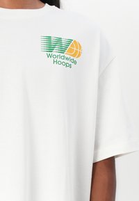 Fehér pamut póló, zöld és sárga "Worldwide Hoops" grafikával, kosárlabda motívummal. Laza szabású, rövid ujjú.