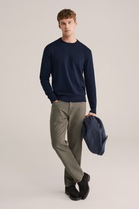 Mornarsko modra dolga majica s krožnim ovratnikom, olivno zelene hlače in temno rjavi semiš čevlji. Drži modro jakno. Nevtralna ozadje.