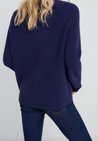 Pull en maille bleu marine avec une coupe ample et des manches larges, featuring une texture côtelée et un ourlet légèrement arrondi. Porté par-dessus un jean foncé.