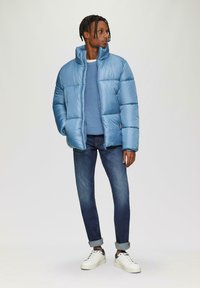 Lichtblauwe pufferjack met een hoge kraag, gedragen over een gestructureerde blauwe trui. Donkere jeans en witte sneakers maken de outfit compleet.