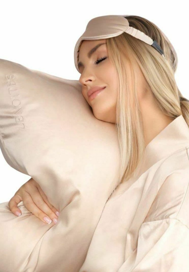 Krämfärgad sidenkudde med präglad logotyp, mjuk textur, hålls nära av en person som bär en matchande sovmask.