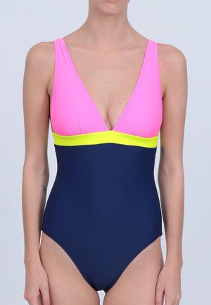 Femme portant un maillot de bain une pièce avec un haut rose, une bande jaune sous la poitrine et un bas bleu marine sur un fond uni.