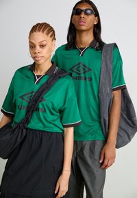 Camisolas de desporto verdes com detalhes pretos e o logótipo "UMBRO" no peito. Ambas as pessoas usam vestuário casual, de corte solto.